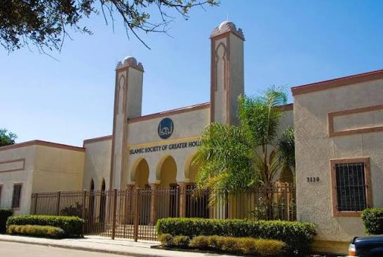 Street view of the masjid, a cream building with two minaret towers, an arched arcade and the Islamic Society of Greater Houston sign above the entrance
